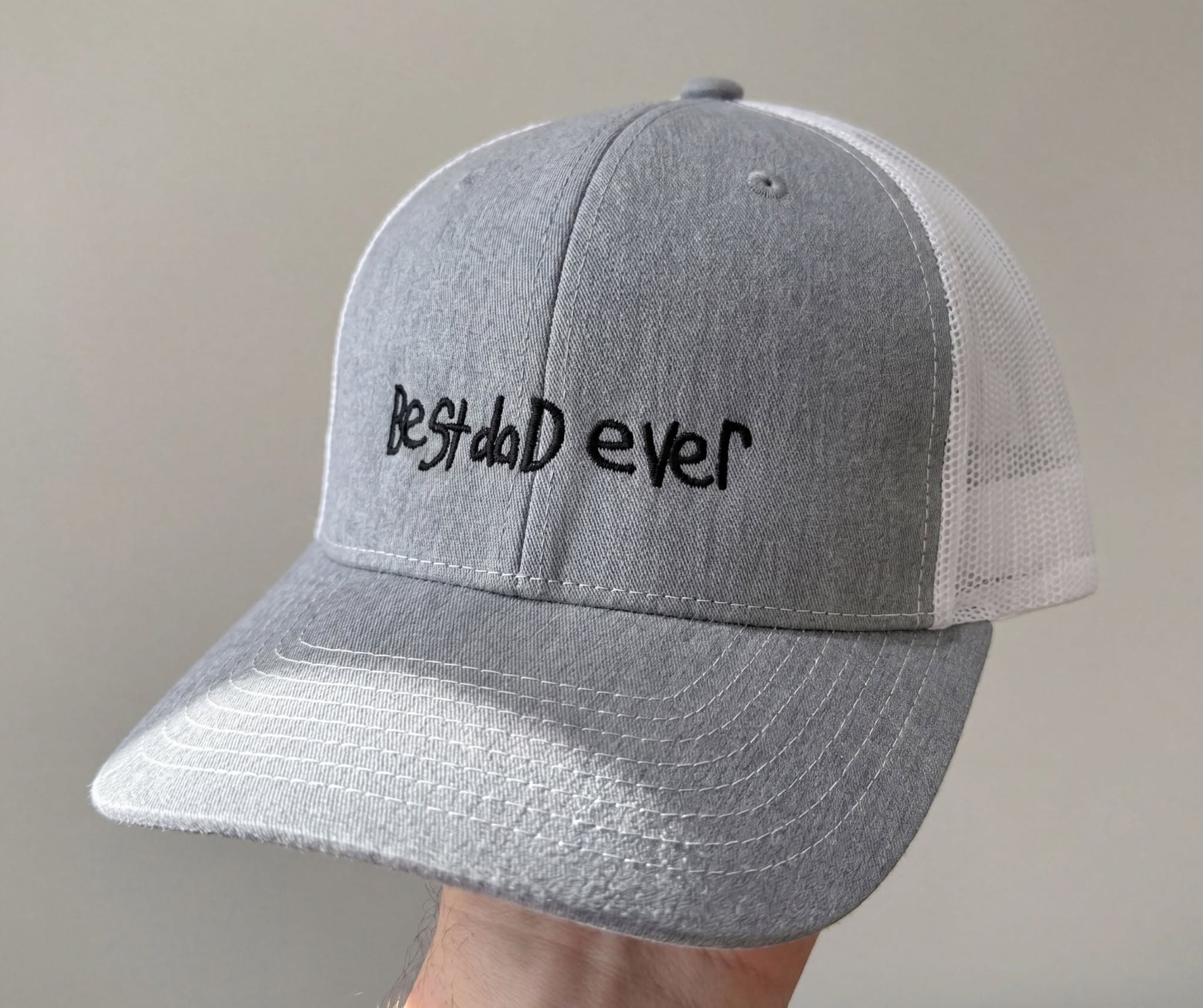 A gray cap embroidered with 'best dad ever' in a child's real handwriting, held up to the camera