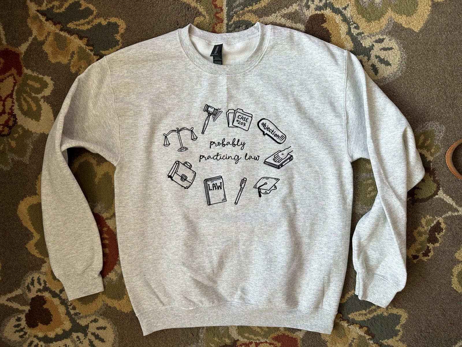 A phone photo of a heather-gray crewneck with a law-themed embroidery design, laid on a patterned living-room rug