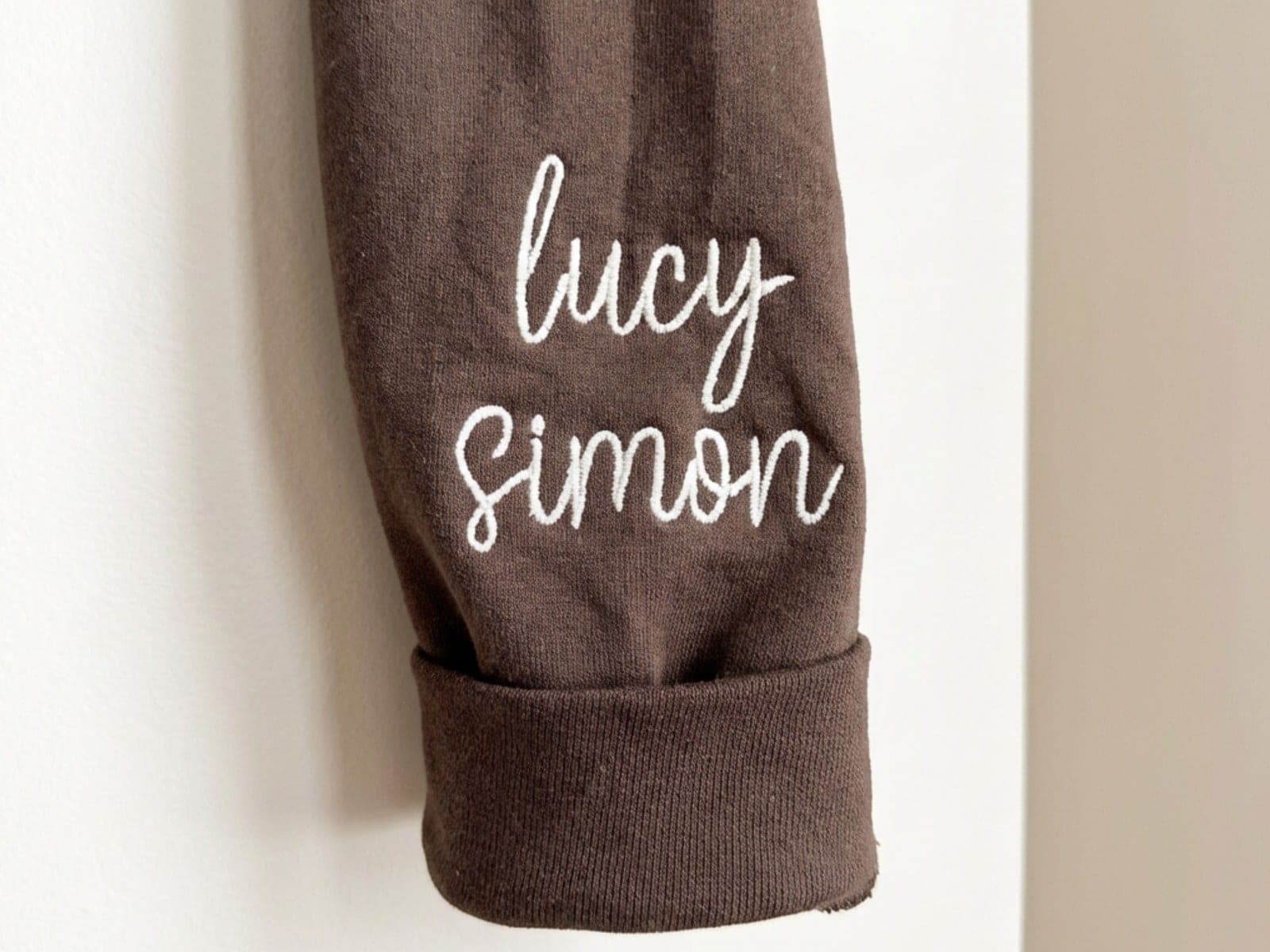 A brown crewneck sleeve with a folded cuff, embroidered with the names lucy and simon in white script