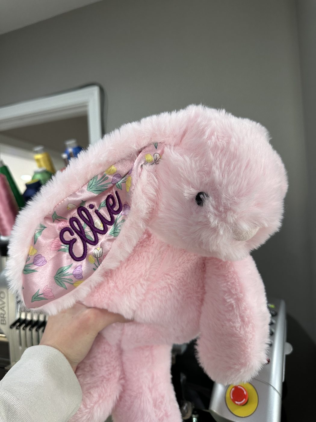 A phone photo of a personalized pink plush bunny held up in the workshop, the machine behind it