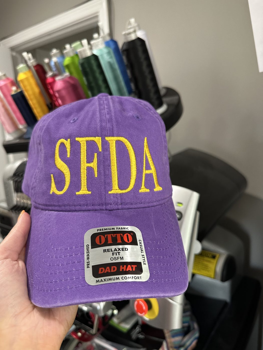 A phone photo of an embroidered purple cap held up in front of the thread rack, size tag still on