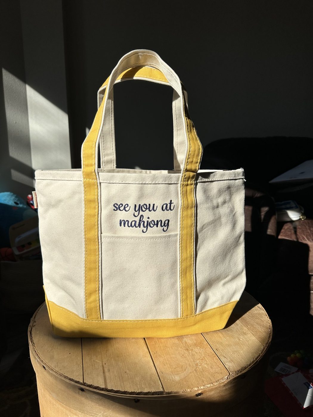 A phone photo of an embroidered canvas tote on a wooden stool in a dim room with harsh window light